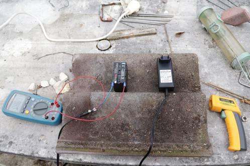 A tile on the table in the backyard, with the NP-F battery, connected to the multimeter and charger, with the temperature gun awaiting