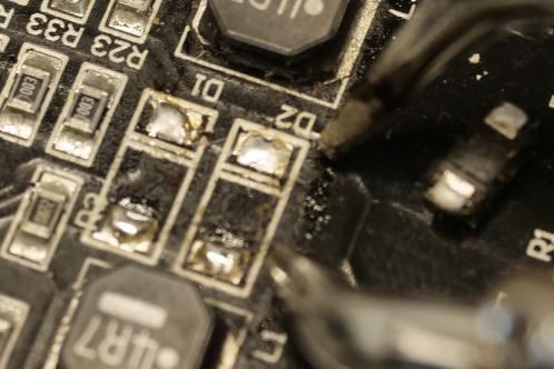 Een zwarte printplaat die de montagelocatie van twee SMD Schottky diodes toont na verwijdering daarvan met het soldeerpincet nog in beeld, bij werkzaamheden aan een Tristar 4 ledlampje