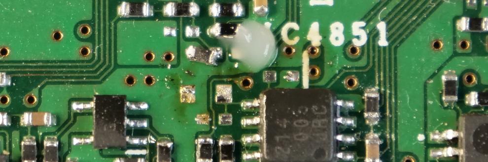 Een klodder witte gel (flux) en een los SMD-weerstandje in 0402 formaat, een SMD IC en diverse andere SMD componenten op een groene printplaat, het analog board van een JVC-GY-HM100 videocamera