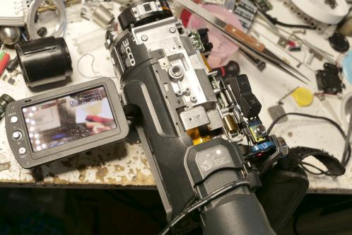 Een aluminium blok met zwarte plastic delen, een uitgeklapt LCD-scherm met beeld: een JVC-GY-HM100 zo ver als nodig samengebouwd om te testen na reparatie