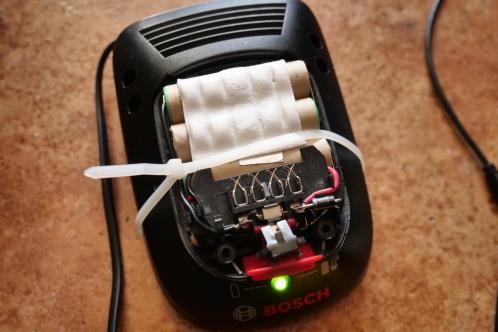 Een zwarte glooiende behuizing van een Bosch PSR acculader, met opengewerkte Bosch PSR lithium schroefmachine-accu gemonteerd, met een witte tie-wrap om deze in bedwang te houden, voor metingen tijdens het laadproces