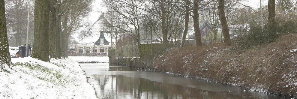 Een waterweg met besneeuwde walkanten met aan het einde een besneeuwd dak van een voormalige stolpboerderij