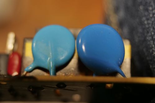 Twee blauwe schijfvormige hoogspanningscondensators die in diameter en dikte verschillen, waarvan 1 gemonteerd is op een elektronicaprint en de andere los ligt