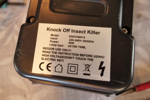 Een zwarte kunststof behuizing met witte identificatieplaat met diverse elektrische symbolen, spanningsvermelding, vermogensopname en waarschuwingen op een vliegenlamp