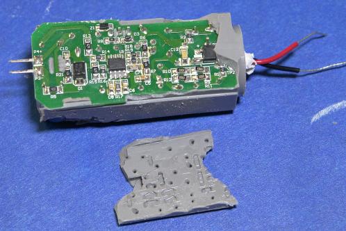 Een groene elektronicaprint met diverse SMD-onderdelen, enkele draden en uitstekende pennen, gedeeltelijk bedekt met grijze gietmassa, waarvan een verwijderd deel onder de print ligt
