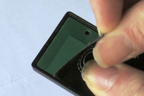 Een groene printplaat in een klein zwart kastje, bedekt door een zwarte sticker die gedeeltelijk losgetrokken is, de radio-afstandsbediening van een Patona DMW-BGGH5RC batterygrip