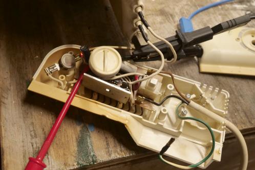 Een witte kunststof behuizing met enige wat bruin verkleurde plekken door hitte toont een bruine elektronicaprint met enkele draden en meet-klemmen aangesloten, ter test van een gerepareerde elektrische kachelthermostaat