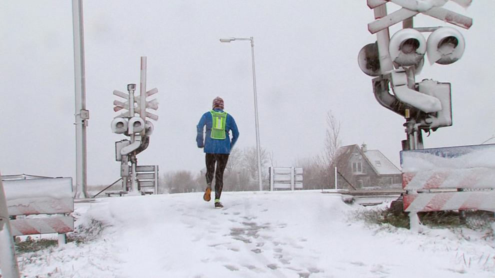 Witte wegen, een overweginstallatie ondergesneeuwd en een hardloper met blauwe jas en groen hesje die temidden van de witte wereld en sneeuwval nog z'n rondje loopt op 10 december 2017
