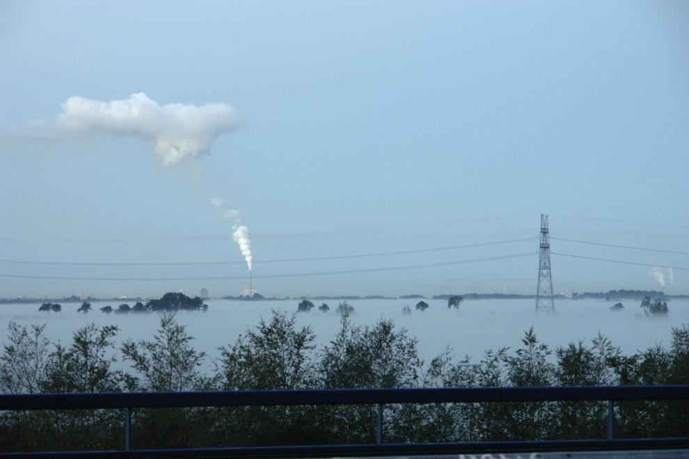 Mistlaag over Purmerland met uitstekende boomtoppen en hoogspanningsmasten, gezien vanaf het 6 meter hoger gelegen Mandelapad te Purmerend. Op de achtergrond vormt rook van de Hemwegcentrale-schoorsteen een nieuwe wolk